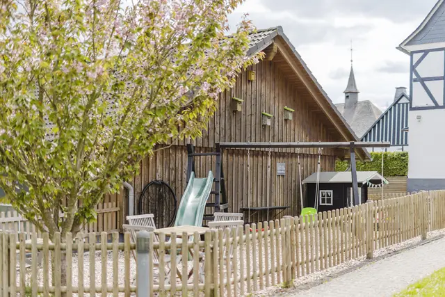 Spielbereich mit Rutsche, Gartenmöbeln und blühendem Baum hinter dem Holzzaun